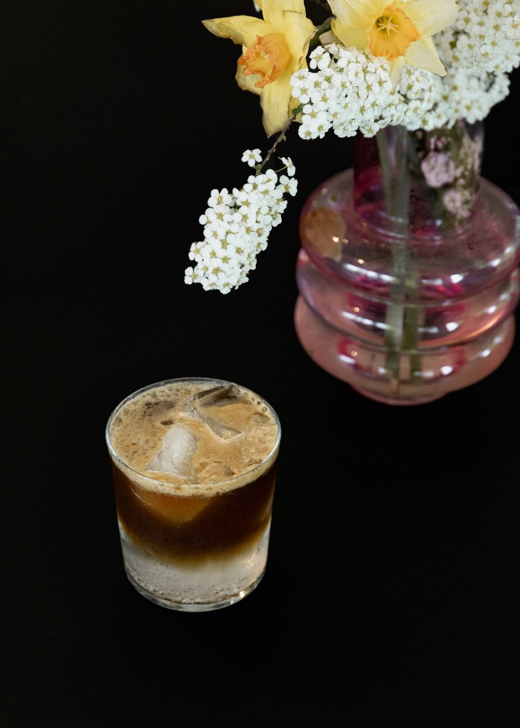 Erfrischendes Eiskaffee-Getränk im Glas neben einer eleganten Blumenvase mit gelben Narzissen auf schwarzem Hintergrund
